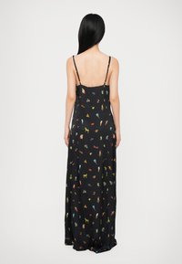 Frau mit langen schwarzen Haaren, die ein schwarzes Maxikleid mit dünnen Trägern und bunten kleinen Mustern trägt, steht mit dem Rücken zu einer weißen Hintergrund.