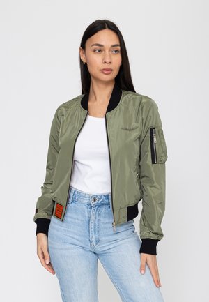 Vrouw die een olijfgroene bomberjack met zwarte boorden en kraag draagt, wit T-shirt en lichtblauwe high-waisted jeans, staand tegen een witte achtergrond.