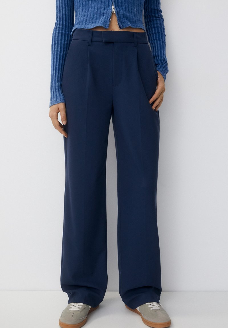 Personne portant un pantalon large bleu marine, un pull court côtelé bleu et des baskets gris clair, debout contre un mur blanc.
