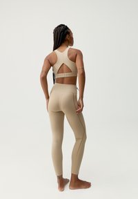 Beige sportovní set obsahuje sportovní podprsenku s výřezem na zádech a odpovídající vysoké legíny s pozlacenými švy. Hladká, elastická tkanina.