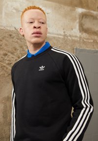 Sweatshirt noir avec design en trois bandes blanches sur les manches, comportant une chemise col bleu en dessous et un logo adidas sur le devant.