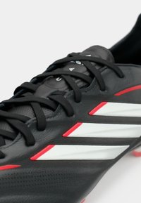 Μαύρο ποδοσφαιρικό παπούτσι με τρεις λευκές ρίγες, κόκκινες λεπτομέρειες, υφή επιφάνειας και μαύρα κορδόνια. Στρογγυλεμένη μύτη και διαμορφωμένες τάπες.