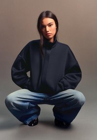 Overmaat donkerblauwe sweatshirt met hoge kraag en geribde manchetten, gecombineerd met losse blauwe spijkerbroek en zwarte schoenen.