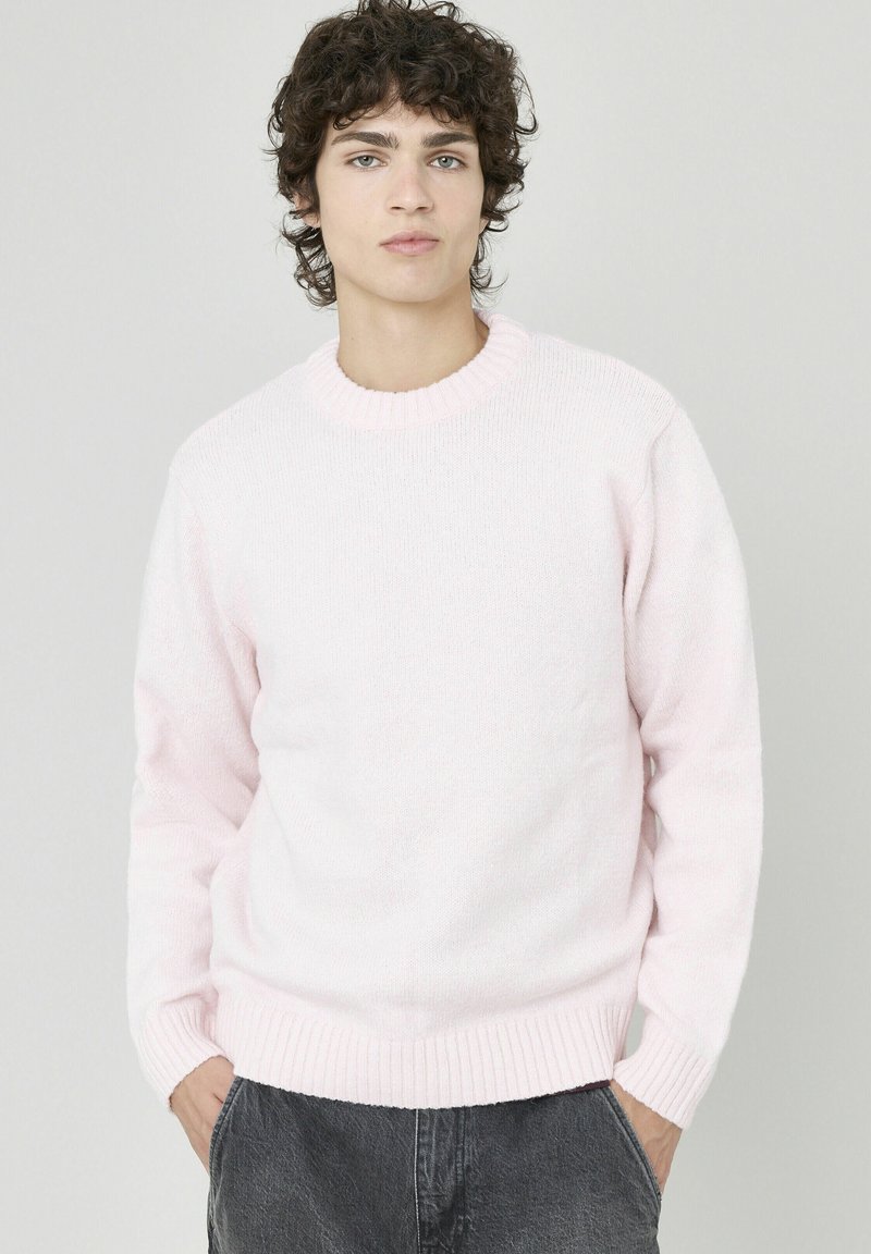 Pull rose clair à col rond avec poignets et ourlet côtelés, fabriqué en matière douce et texturée. Porté avec un pantalon gris foncé.