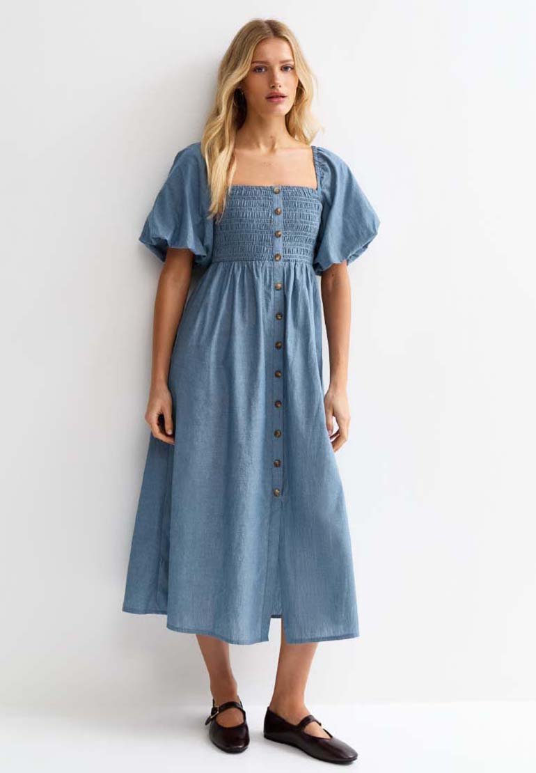 Woman standing wearing a blue midi dress with puff sleeves, front buttons, square neckline, and black Mary Jane shoes.