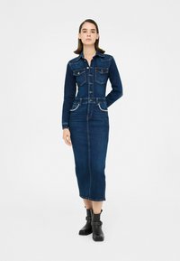 Abito di jeans con design a collo, colore blu scuro, due tasche frontali, dettagli con borchie sulle tasche e silhouette aderente, abbinato a stivali neri.