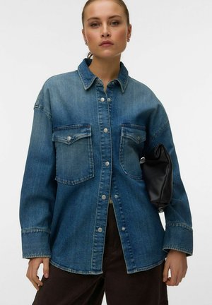 Femme portant une veste en jean bleu oversize avec des boutons-pression et deux poches poitrine, tenant une pochette en cuir noire sous son bras.