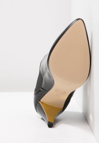 Chaussure noire à talons hauts avec un bout pointu, présentant une finition brillante en vernis et un tissu noir texturé sur les côtés. La semelle est nude avec de fines rainures.