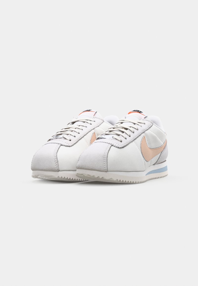 Baskets blanches avec une tige en cuir suédé et en cuir, arborant un swoosh rose clair, des accents gris, un devant à lacets et une semelle en caoutchouc texturé.
