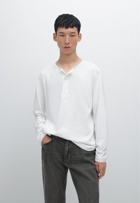 Langärmliges, weißes Henley-Shirt mit lockerer Passform, V-Ausschnitt und drei Knöpfen, kombiniert mit dunkelgrauen Jeans.