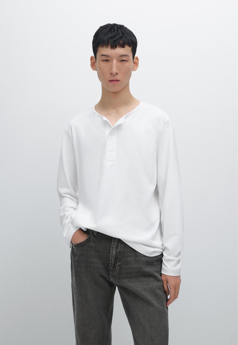 Langärmliges, weißes Henley-Shirt mit lockerer Passform, V-Ausschnitt und drei Knöpfen, kombiniert mit dunkelgrauen Jeans.