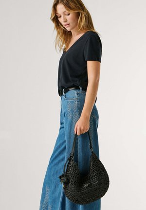 Woman with light brown hair wearing a black V-neck shirt and high-waisted blue jeans, holding a black woven shoulder bag.