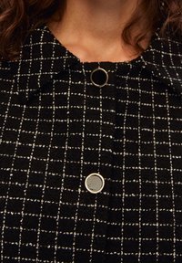 Zwart-wit geruit stof met een gestructureerde afwerking, voorzien van een kraag en twee ronde knopen—een metalen, een met stof bekleed.
