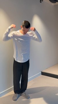 Άνδρας που ρυθμίζει το λευκό πουκάμισο με μακριά μανίκια, στέκεται σε εσωτερικό χώρο δίπλα σε γκρι τοίχο κοντά σε σκάλες, φοράει μαύρο παντελόνι και γκρι παντόφλες.