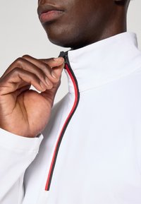 Jachetă albă cu fermoar negru și roșu. Gulerul este înalt, iar o mână ajustează fermoarul. Textură netedă, design modern.