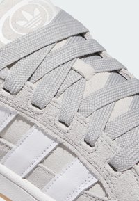 Grå semsket sneaker med hvite striper, vevde grå skolisser og et rundt logo-patch på tungen med trebladdesign.
