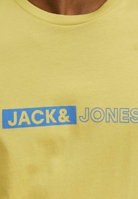 Camiseta de algodón amarilla con cuello redondo, con letras en bloque azules que dicen "JACK&JONES" en el frente. Textura suave y diseño informal.