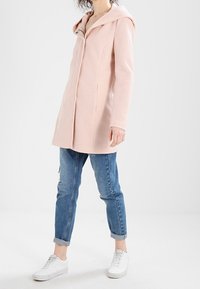 Abrigo con capucha de color rosa claro y corte entallado, con cierre frontal, combinado con jeans azules arremangados y zapatillas blancas. Textura suave.