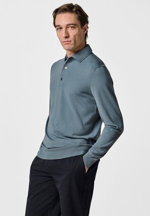 Mann in einem taubenblauen langärmeligen Poloshirt und schwarzen Hosen, der mit einer Hand in der Tasche vor einem schlichten Hintergrund steht.