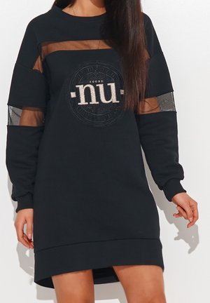 Kvinde iført sort langærmet kjole med gennemsigtige meshpaneler på ærmer og bryst, med et "nu"-emblem foran.