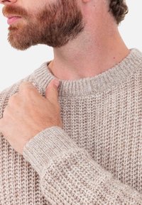 Beiger gestrickter Pullover mit rundem Ausschnitt, gerippten Bündchen und strukturiertem Muster. Nahaufnahme zeigt eine Hand, die den Schulterbereich berührt.