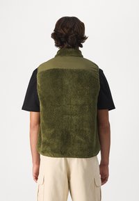 Green fleece vest with a plush texture, contrasting olive nylon upper, high collar, and sleeveless design, worn over a black shirt.