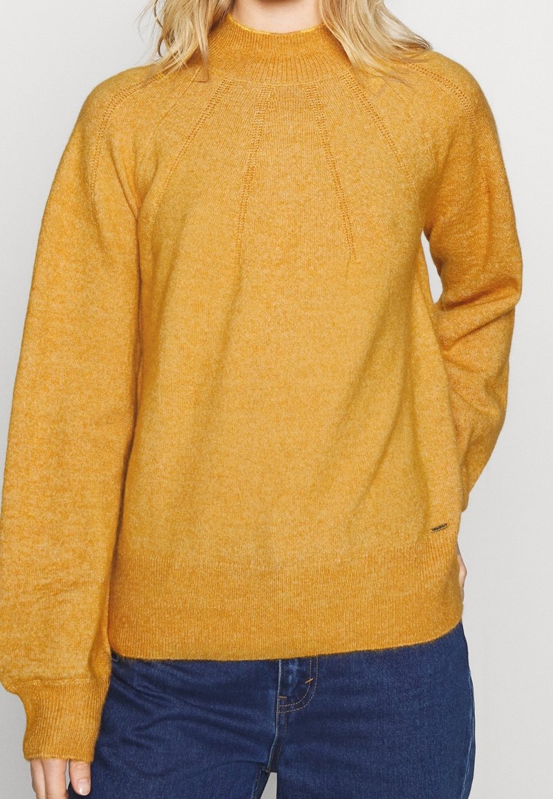 Woman wearing a mustard yellow knit sweater with long sleeves and high neckline, paired with blue denim jeans against a plain background.