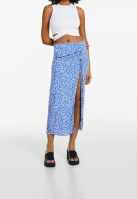 Modrá zavinovací sukně s bílým květinovým vzorem, s bočním rozparkem. Spárována s bílým topem bez rukávů a černými platformovými sandály.