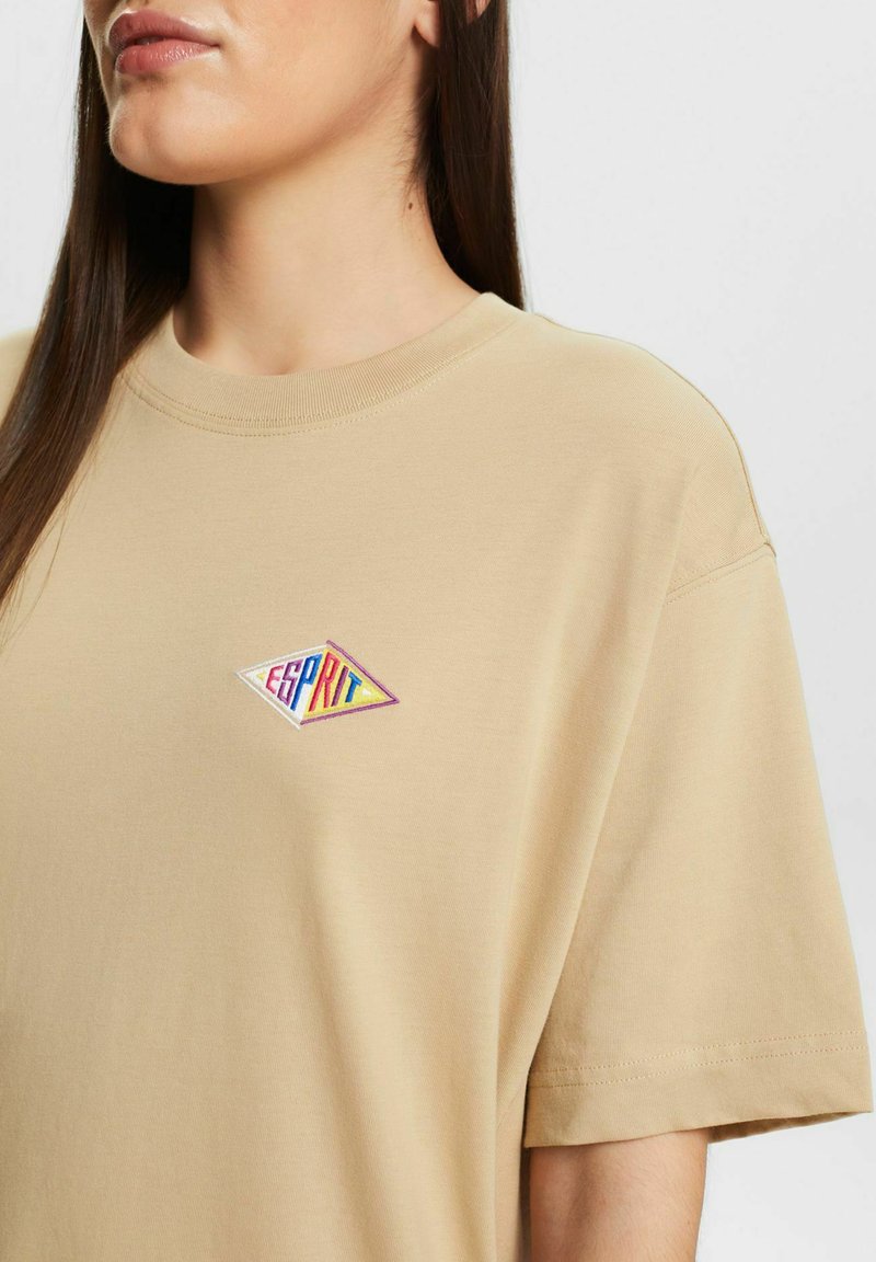 Kobieta w beżowej koszulce z krótkim rękawem, z kolorowym diamentowym logo "Esprit" na piersi, skrócona przy szyi i ramionach.