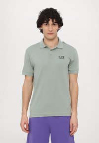 Svetlozelená polo košeľa s krátkymi rukávmi, s rebrovaným golierom a čiernym logom EA7 na ľavej hrudi. Hladká bavlnená textúra.