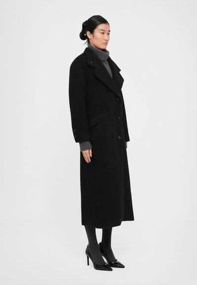Femme debout, portant un long manteau noir à boutons, un pull col roulé gris, des collants gris et des chaussures à talons hauts noires sur fond blanc.