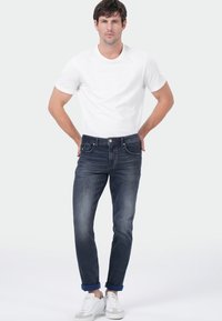 Man staat met zijn handen in zijn achterzakken, draagt een wit T-shirt, donkere opgerolde jeans en witte sneakers voor een effen achtergrond.