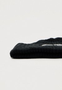 Black knitted beanie, ribbed texture, folded design. Features white logo patch with text on the side. Soft, warm material.
