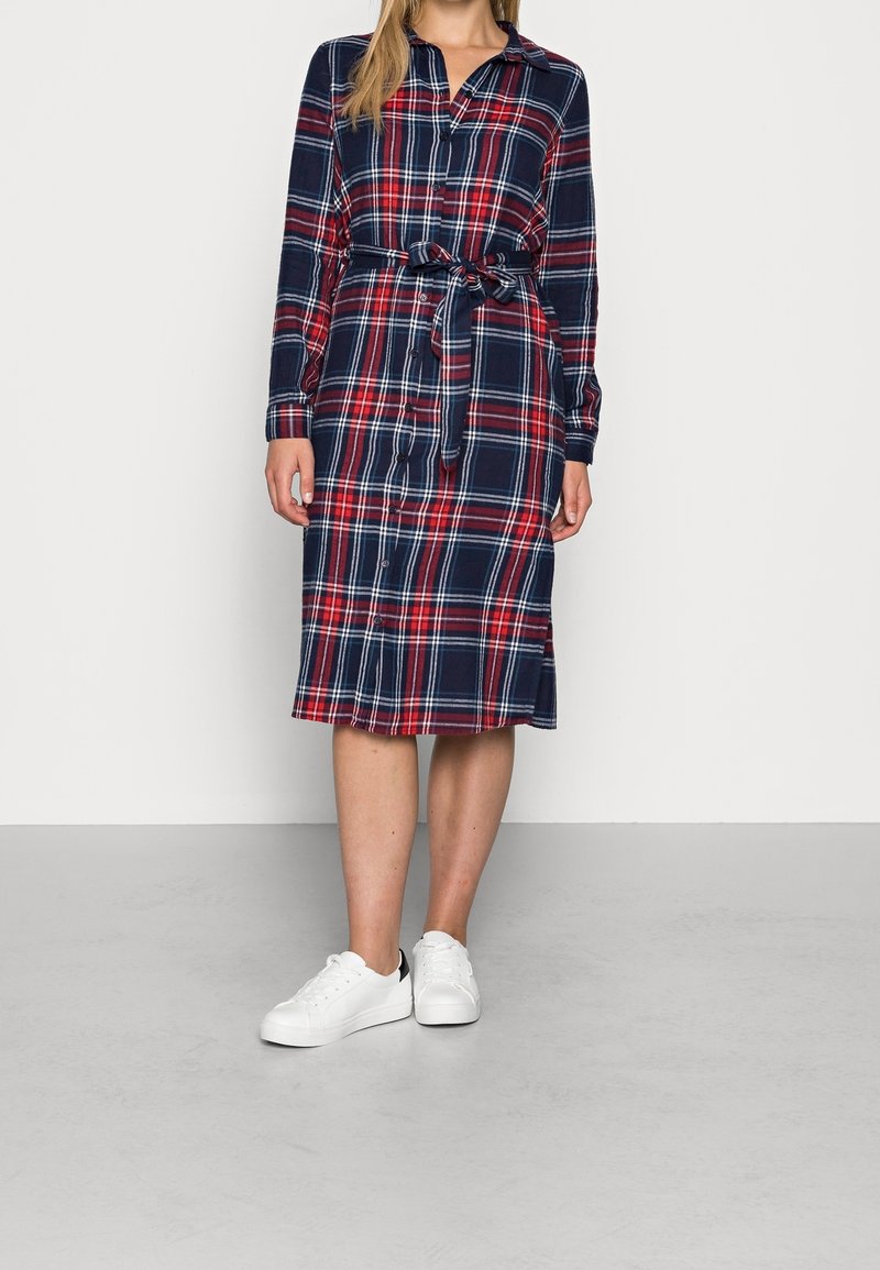 Abito camicia a quadri in blu navy, rosso e bianco. Realizzato in tessuto morbido, presenta un colletto, chiusura con bottoni e una cintura legata in vita. Indossato con sneakers bianche.