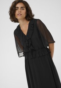 Robe noire avec un décolleté en V froncé et de courtes manches transparentes, taille cintrée avec un ourlet froncé, et jupe fluide avec des plis.