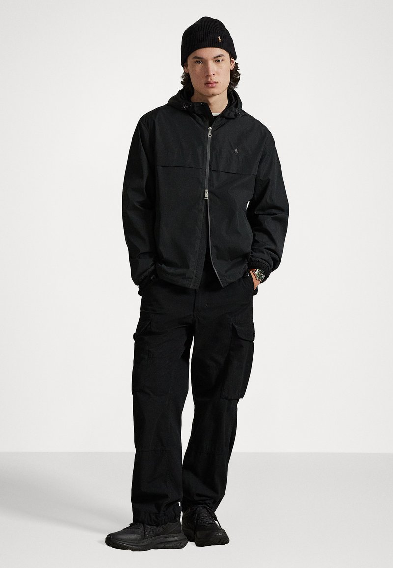 Veste à capuche noire avec fermeture éclair à l'avant, associée à un pantalon cargo noir avec poches latérales. Bonnet en tricot texturé et baskets noires.