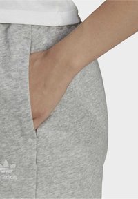 Ruka v kapse šedých tepláků Adidas s viditelným logem, nahoře s bílým tričkem.