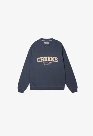 Sweatshirt bleu marine à manches longues, avec des poignets côtelés et un col ras du cou. Présente l'inscription 'CREEKS ORIGINAL Depuis 1974' en beige, imprimée sur le devant.
