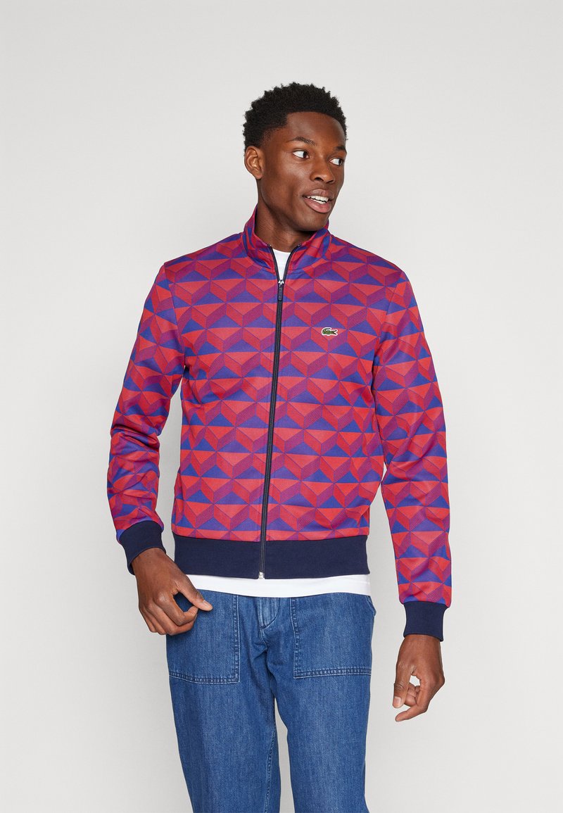 Veste zippée pour homme en violet et rouge avec un motif géométrique. Dispose d'un col haut et de poignets côtelés. Logo Lacoste sur la poitrine gauche.