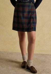 Spódnica w kratę w odcieniach ciemnego niebieskiego, zielonego i czerwonego; długości do kolan, z płaskim pasem i subtelnym połyskiem. Zestawiona z brązowymi mokasynami i beżowymi skarpetkami.