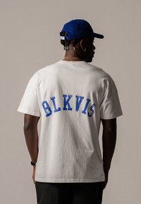 T-shirt en coton blanc avec un texte "BLKVIS" bleu de style vintage au dos. Manches courtes, coupe décontractée et texture lisse. Détails supplémentaires de la casquette visibles.