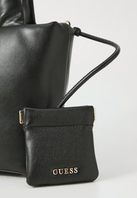 Zwarte leren Guess handtas met een kleine bijbehorende tas, voorzien van een gouden logo en sluiting, tegen een effen lichte achtergrond.