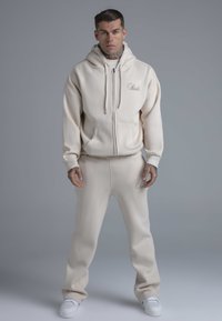 Beiger Kapuzenpullover mit Reißverschluss und Kängurutaschen, kombiniert mit passenden Jogginghosen. Der Stoff wirkt weich und strukturiert und weist dezente Logoakzente auf.