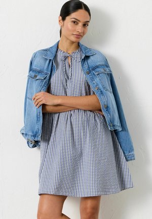 Frau mit dunklem Haar in einem tiefen Dutt trägt ein blaues Karo-Kleid mit Bindekragen und eine hellblaue Jeansjacke, die über ihre Schultern gelegt ist.