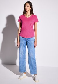 Růžové tričko s krátkým rukávem a výstřihem do V v kombinaci s světle modrými širokými džíny a bílými teniskami. Tričko má jemnou texturu a volný střih.