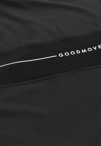 Camiseta deportiva negra con una banda texturizada negra que lleva el texto "GOODMOVE" en blanco y detalles perforados. Tejido suave con un diseño elegante.