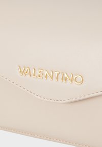Krmová kožená kabelka s trojúhelníkovým klopem, s pozlaceným embosovaným logem "VALENTINO" a kontrastním prošíváním podél okrajů.