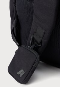 Mochila negra de tela con un panel de malla texturizada; cuenta con un pequeño bolsillo con cremallera adjunto por un clip, que muestra un logo sutil.