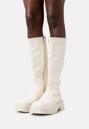 Botas blancas hasta la rodilla hechas de cuero liso, con cremallera lateral, suela de goma gruesa con textura acanalada y diseño de punta redondeada.