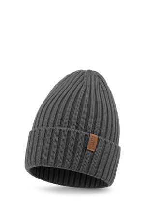 Homme portant un bonnet en maille côtelée gris foncé et une veste noire avec un léger sourire, face à un fond clair uni.